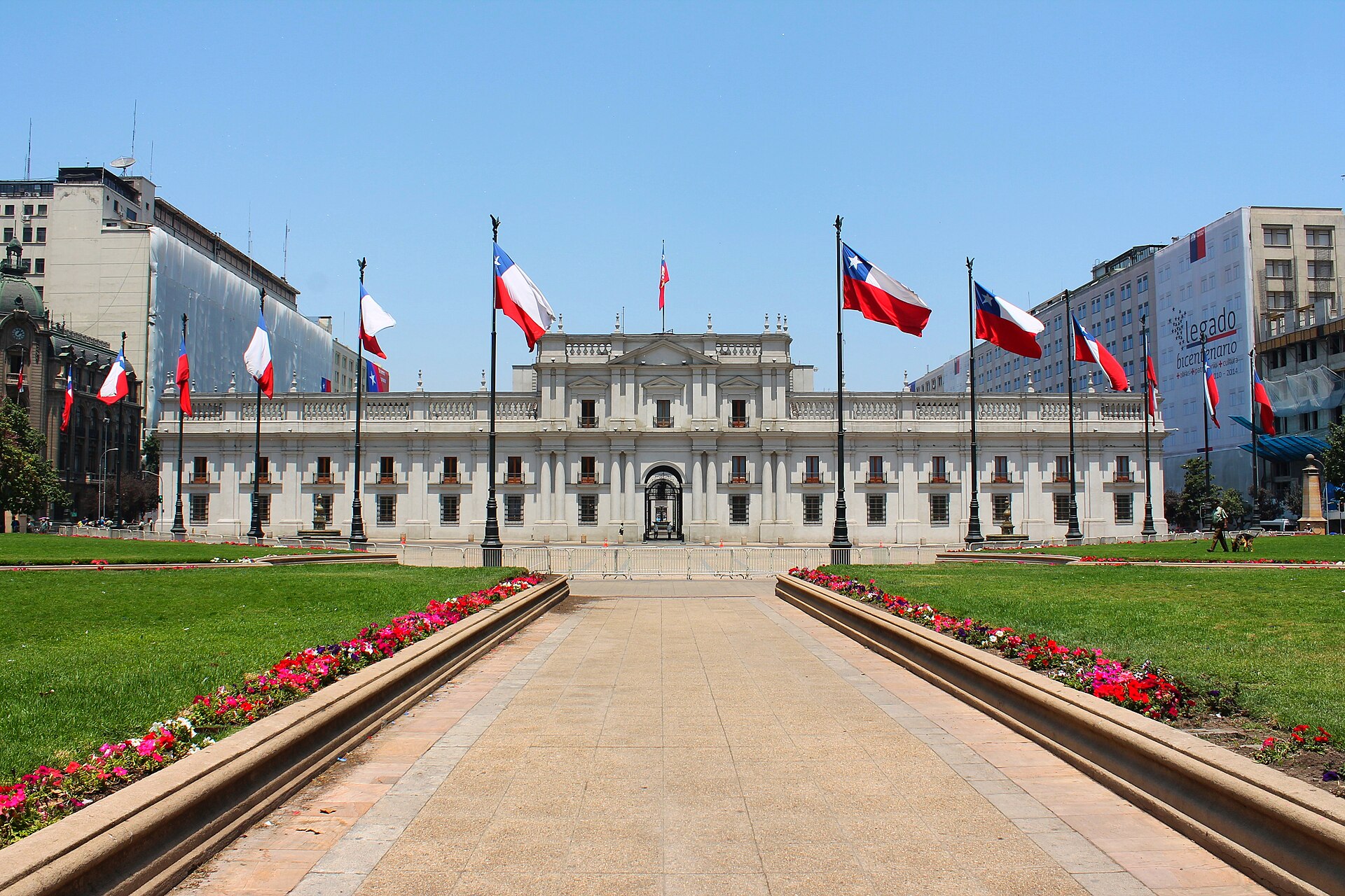La Moneda Palace in Santiago, Chile, with Chilean flags above a flower-lined plaza on a sunny day.