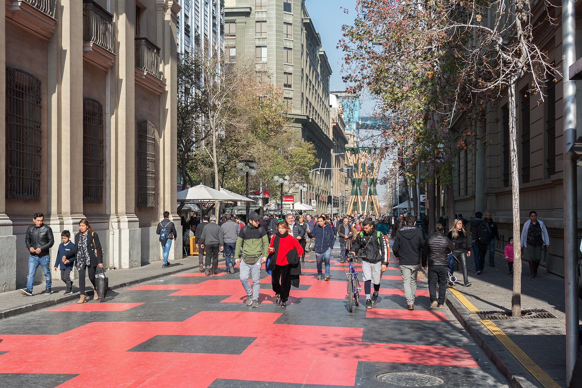 Pedestrians and a cyclist walk along a red-and-black patterned street lined with historic buildings and autumn trees.