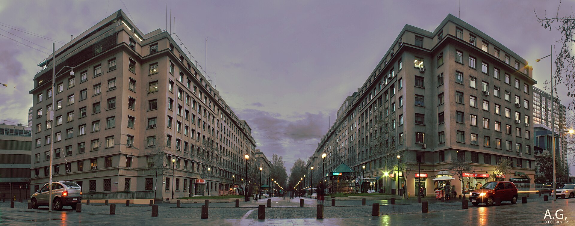 Panoramic dusk view of a pedestrian boulevard between apartment buildings, with streetlights, shops, and cars on wet pavement.