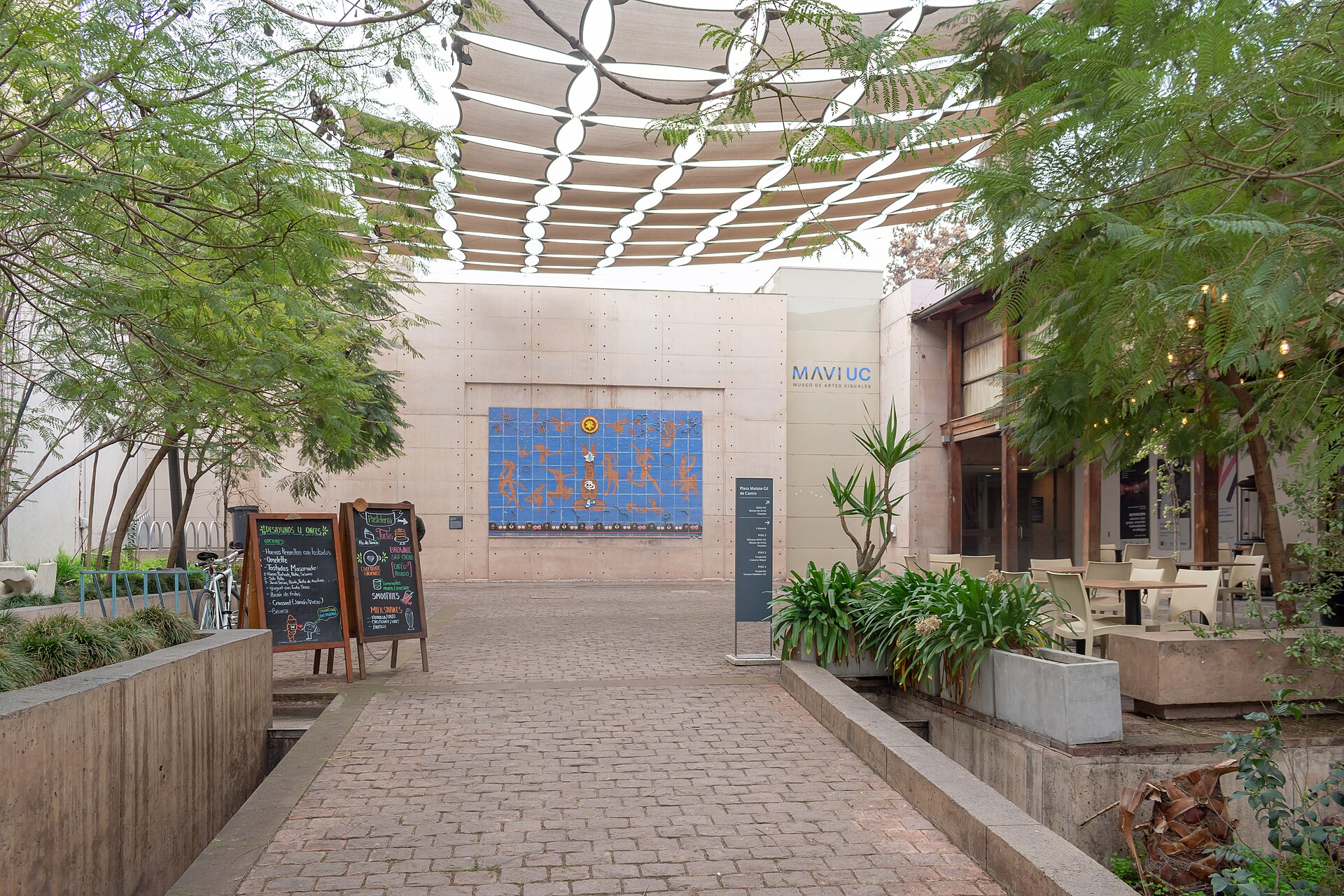 Tree-lined courtyard entrance to MAVI UC art museum, with overhead shade canopy, wall mural, café tables, and menu boards.