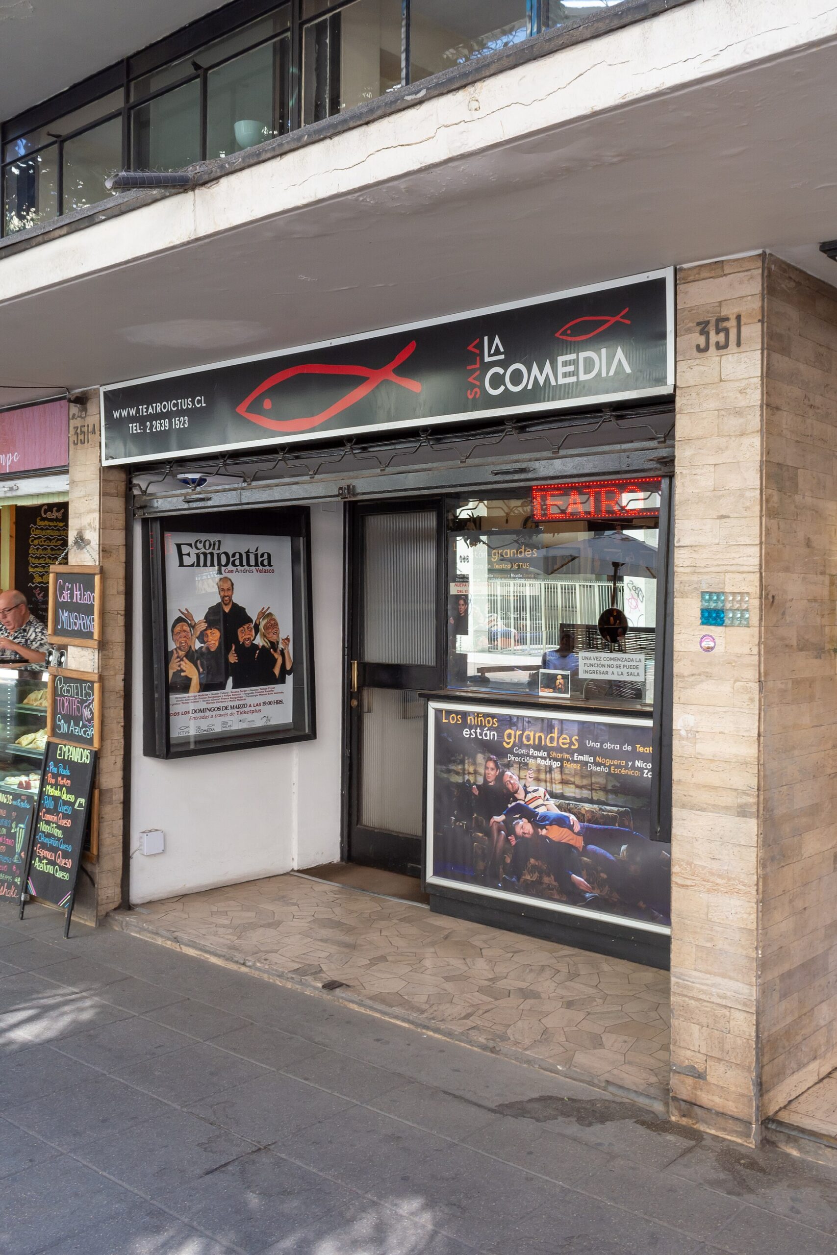 Street-level view of Teatro Ictus entrance with comedy sign, theater posters, and neighboring café menu boards.