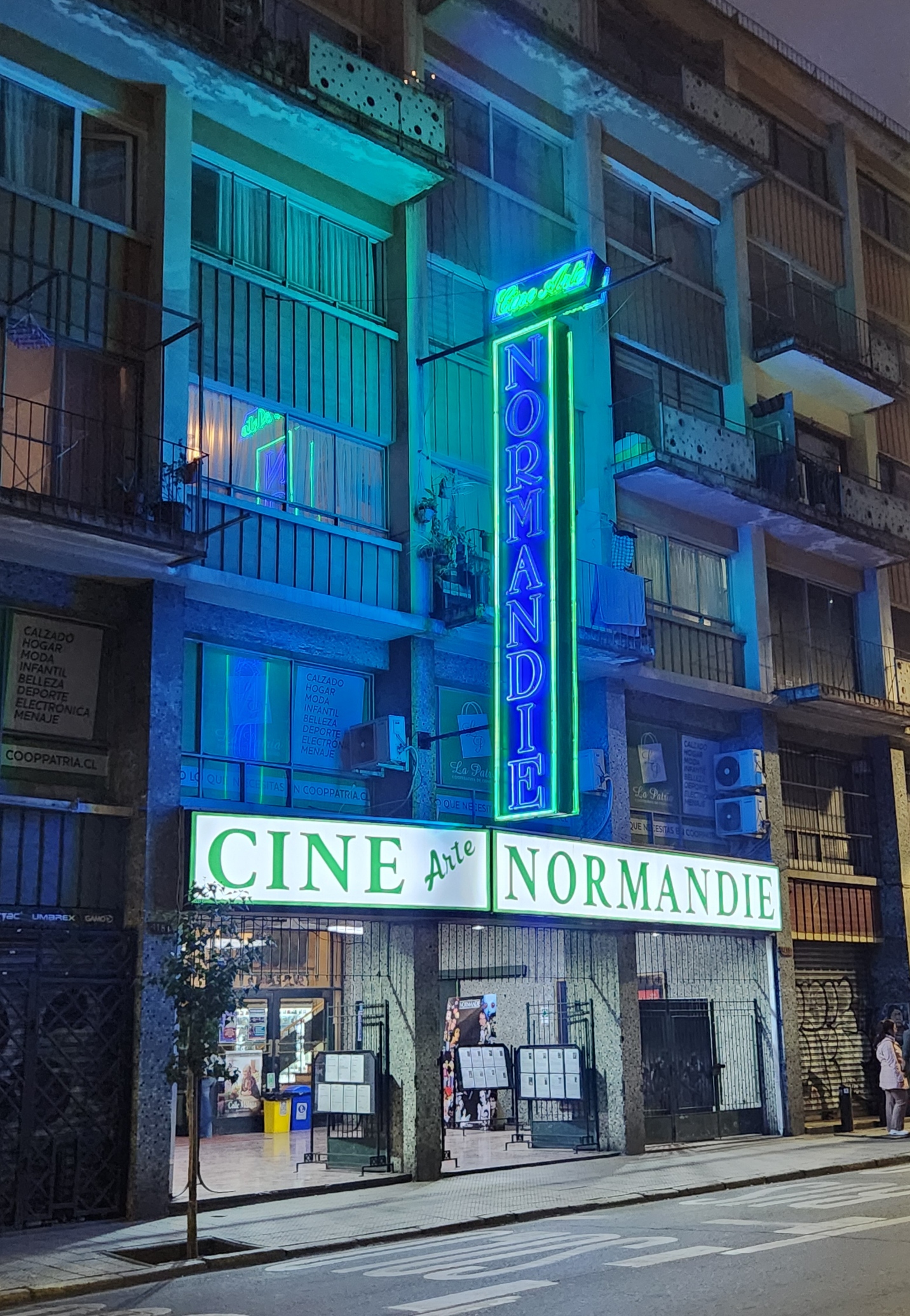 Neon-lit Cine Arte Normandie cinema entrance beneath a tall apartment building on a city street at night.