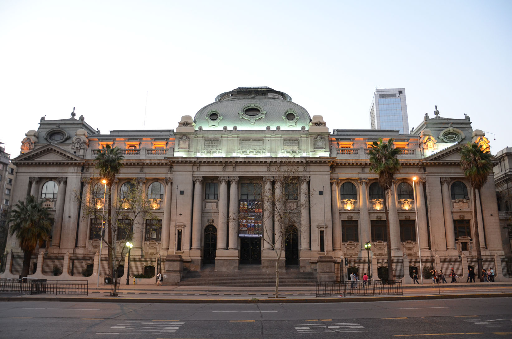 Ornate neoclassical building with a domed center illuminated in green and orange at dusk, with pedestrians and palm trees outside.
