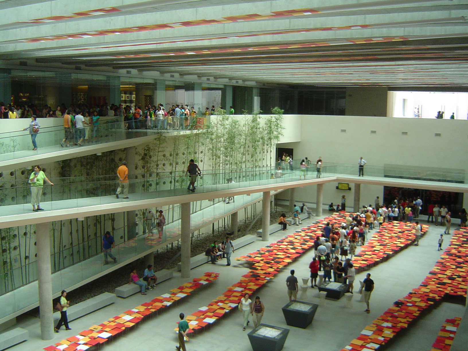 Visitors walk through a modern museum atrium displaying long rows of colorful books or tiles on the floor.