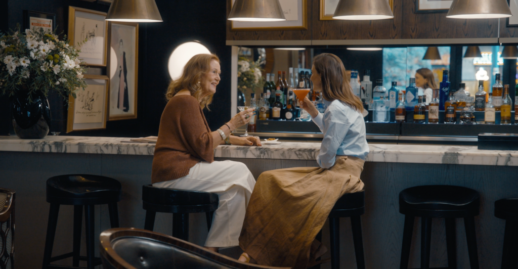 Two women smile and raise cocktails while chatting at a warmly lit bar lined with bottles.