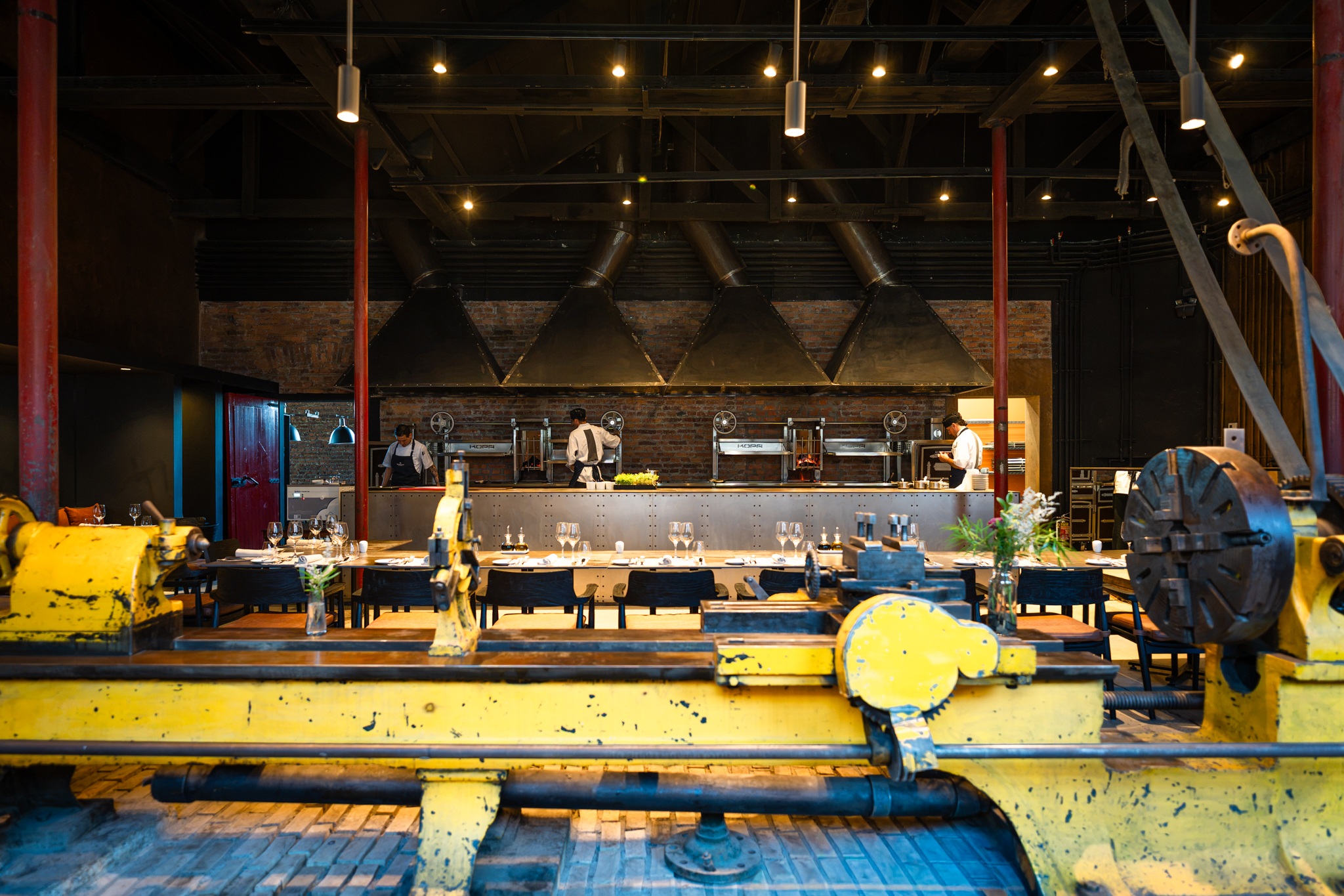 Chefs prepare food in an industrial-style restaurant kitchen with exposed brick, metal hoods, and yellow machinery in the foreground.
