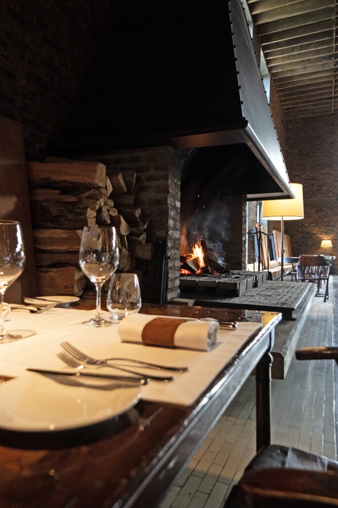 Elegant restaurant table set beside a large stone fireplace, with stacked logs, wine glasses, and warm lamplight.