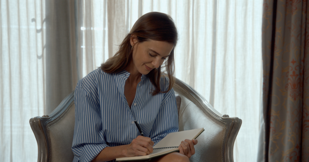 Woman in a blue striped shirt writes in a notebook while seated in an upholstered armchair by a bright window.