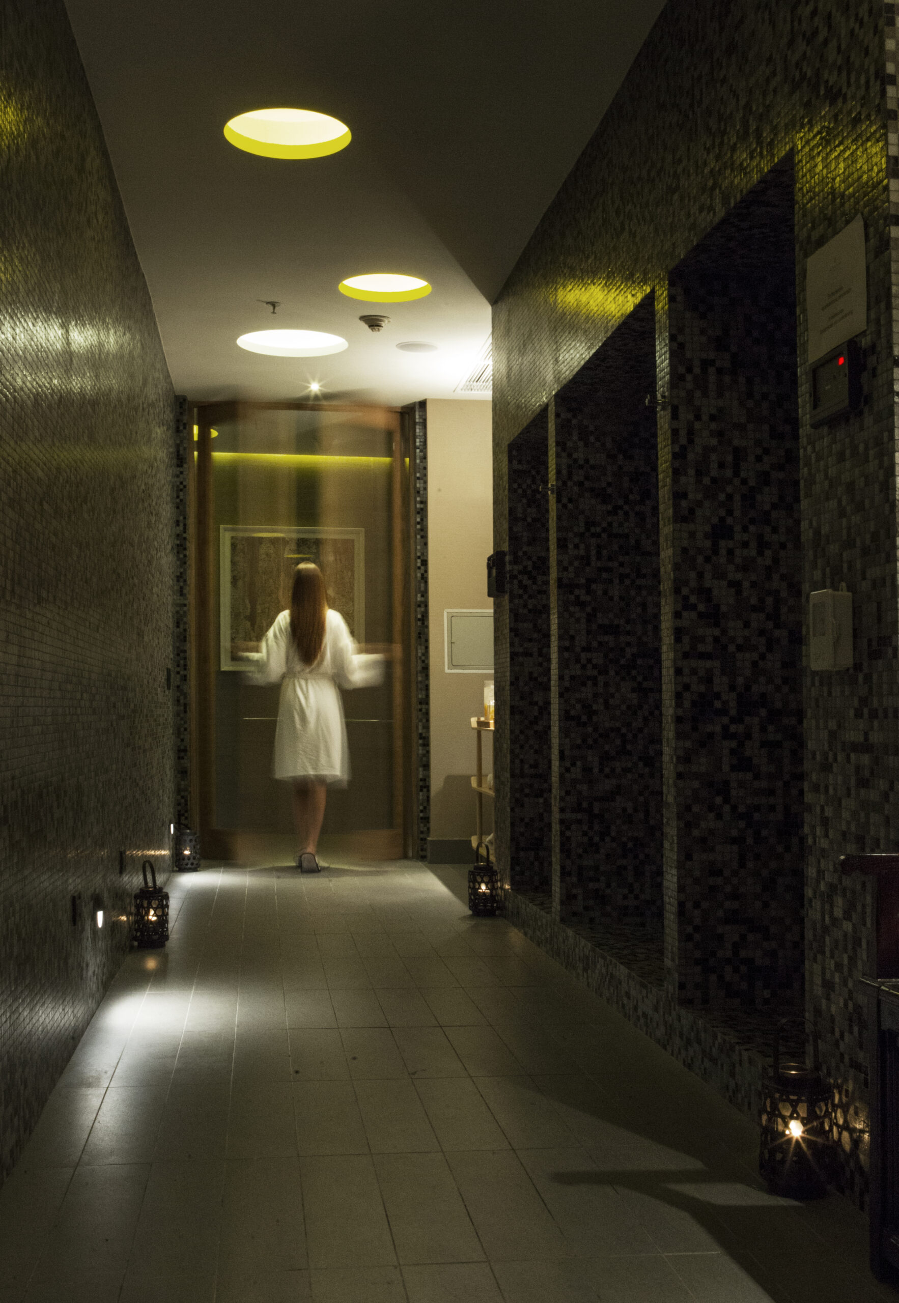 A woman in a white robe walks down a dim, tiled spa corridor lit by lanterns and warm ceiling lights.