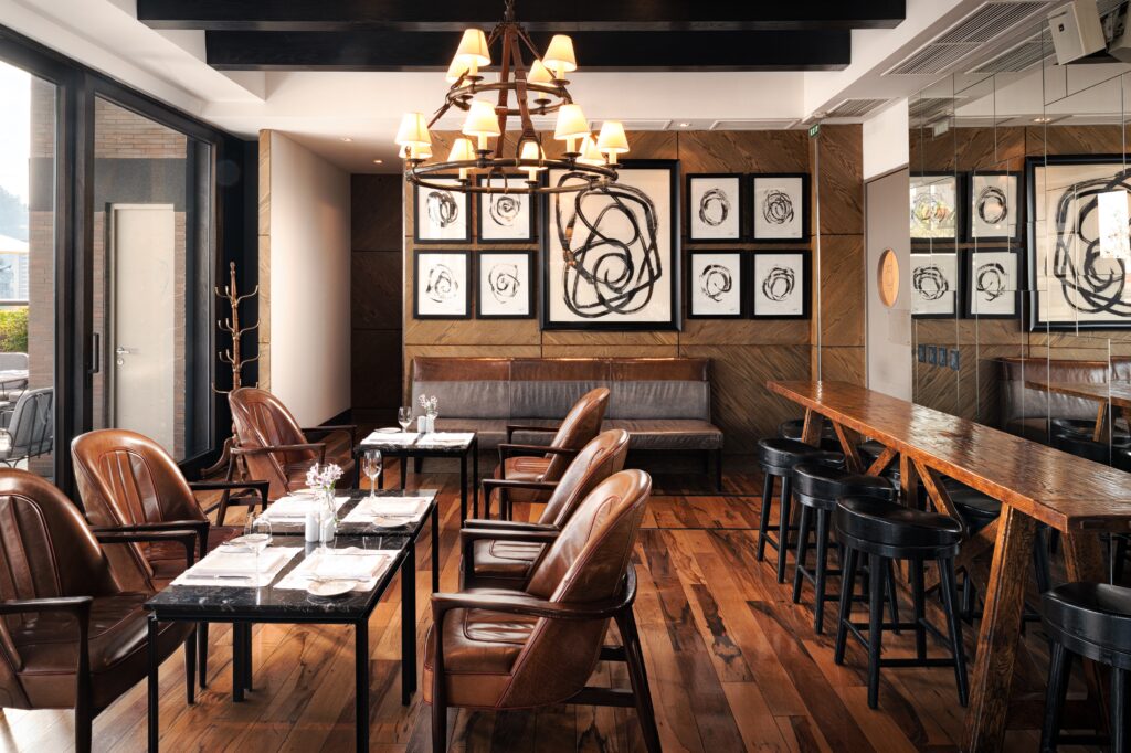 Upscale restaurant interior with leather chairs, set dining tables, wood-paneled walls, abstract art, and a glowing chandelier.