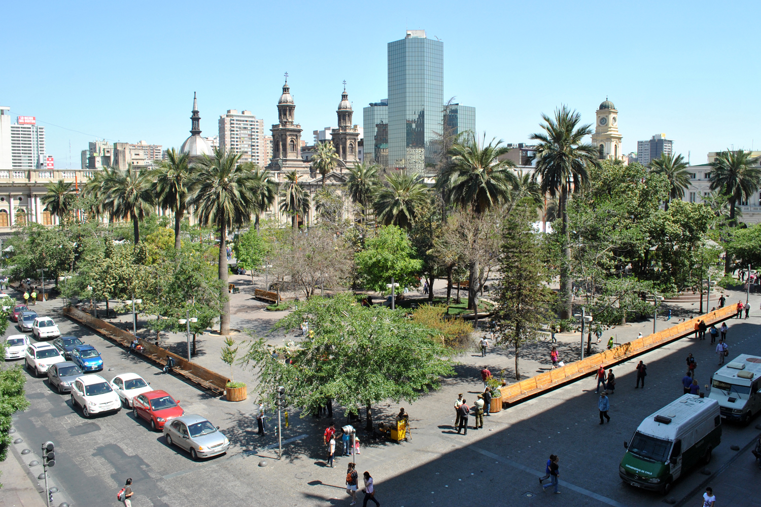 Santiago, Chile city square with palm trees, pedestrians, parked cars, and historic church towers beneath modern skyscrapers.