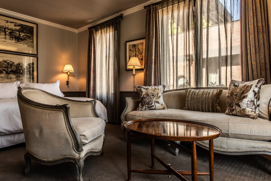Elegant hotel suite with a bed, upholstered sofa and armchair, oval coffee table, framed art, and sunlit draped windows.