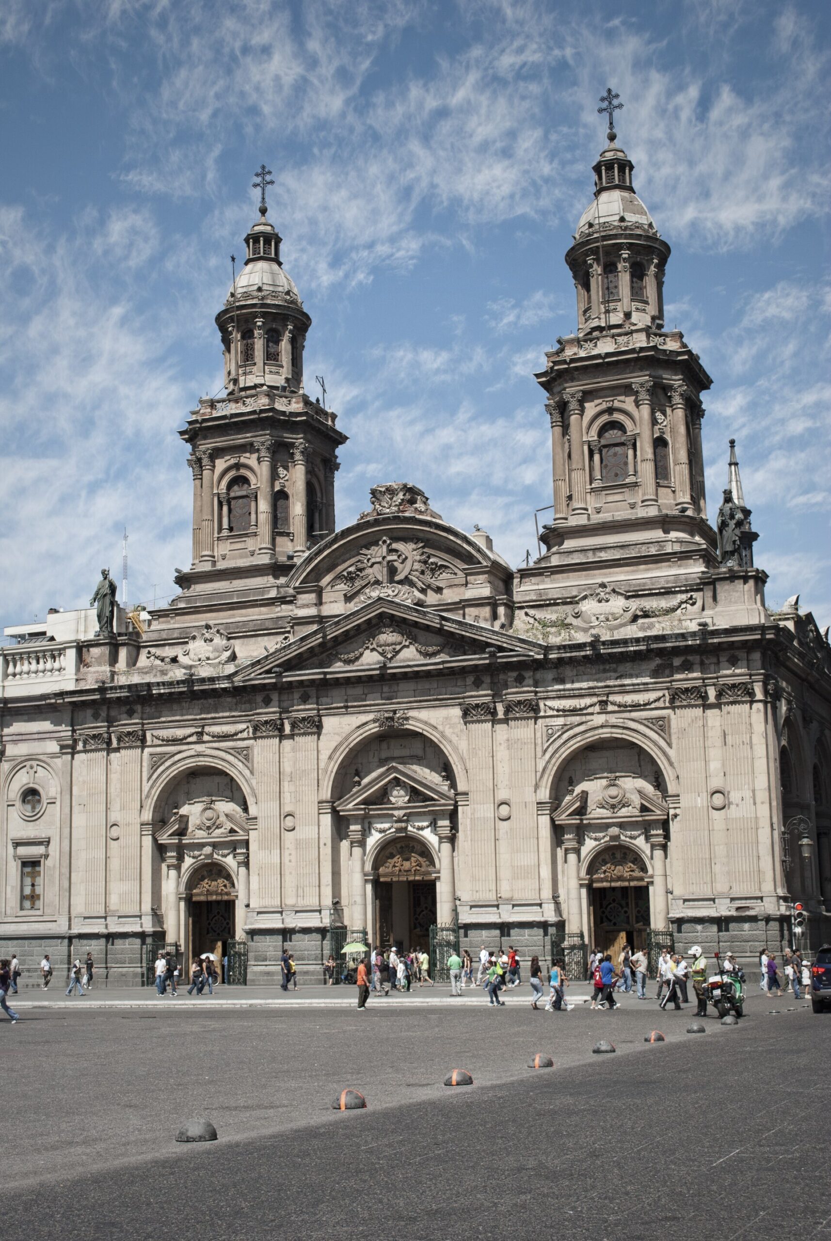 Ornate stone cathedral with twin bell towers, people crossing the plaza, and a blue, cloud-streaked sky.