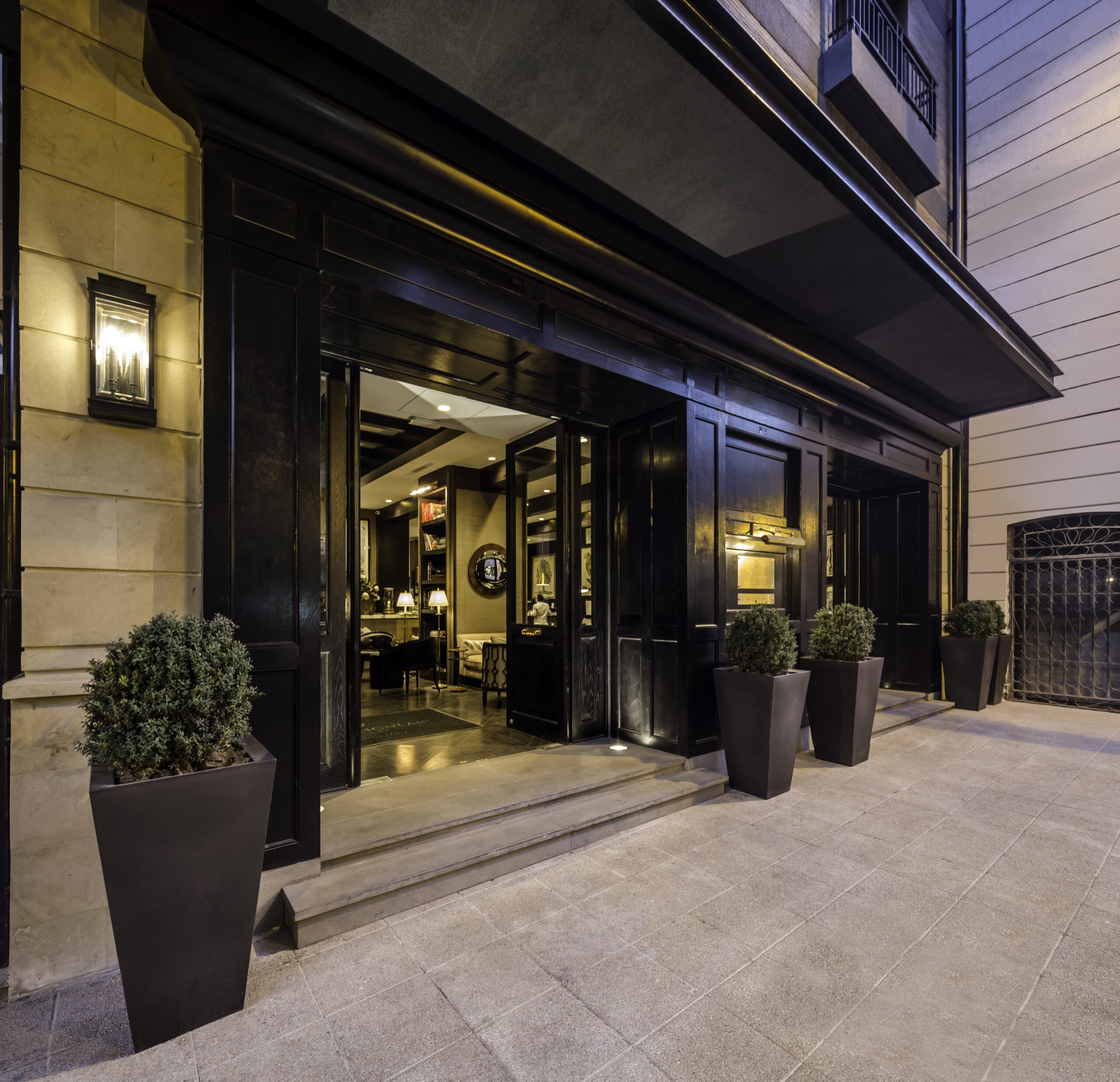 Elegante entrada del hotel por la noche con un dosel negro curvo, vestíbulo iluminado, escalones de piedra y arbustos en maceta.