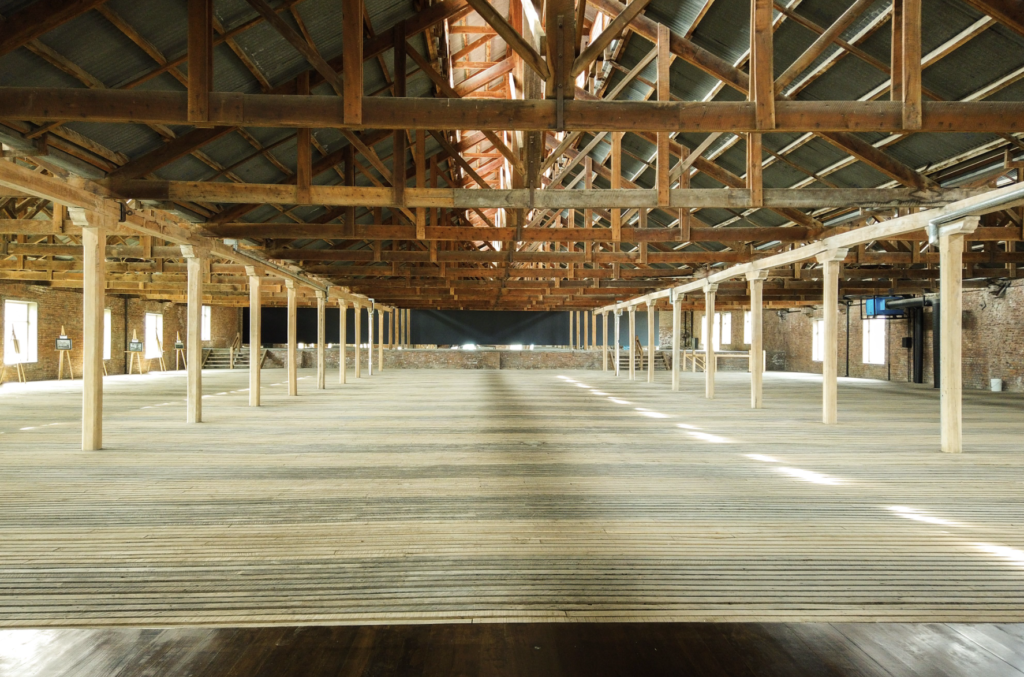 Vast empty warehouse interior with timber roof trusses, brick walls, wooden columns, and sunlight across the plank floor.