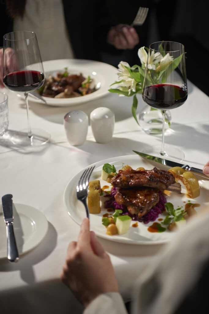 Elegant restaurant dinner with plated grilled meat, red wine glasses, white flowers, and diners seated at a table.