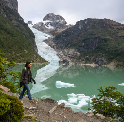 Hiker walks beside a turquoise glacial lake with floating ice, backed by a glacier and rugged mountains in Patagonia.