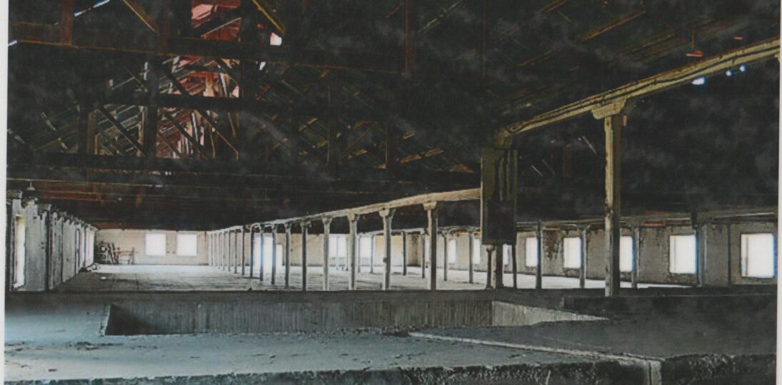 Interior of a large industrial building with exposed steel roof trusses, support columns, and rows of bright windows.