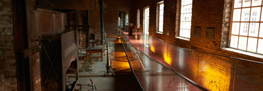 A glass walkway runs through a historic brick factory interior with old machinery, exposed beams, and tall windows.
