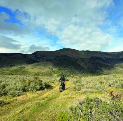 A mountain biker rides a grassy trail through rolling green hills beneath a cloudy blue sky.