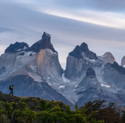 Amanecer en Pehoe (9 de 1) Hiker on a forested ridge overlooks the jagged granite peaks of Torres del Paine beneath a cloudy sky.