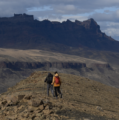 Two hikers with backpacks and trekking poles cross a rocky ridge beneath a dramatic mountain plateau and cloudy sky.