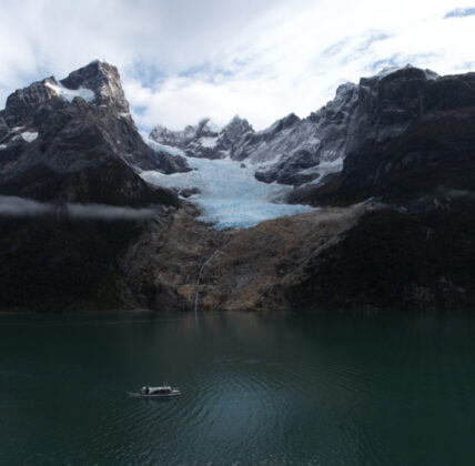 Small boat on a turquoise glacial lake beneath a glacier and rugged, snow-covered mountains in Patagonia.