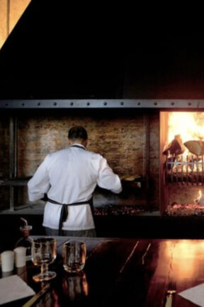 Screenshot Chef in white jacket cooks beside a blazing open-fire grill in a rustic restaurant kitchen, with a dining table in the foreground.