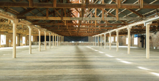 Screenshot Empty historic warehouse interior with exposed wooden roof trusses, brick walls, timber posts, and a wide plank floor.