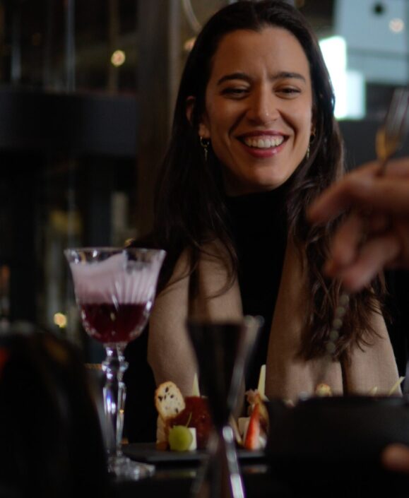 Smiling woman enjoys cocktails and appetizers with a companion at a dimly lit restaurant bar.