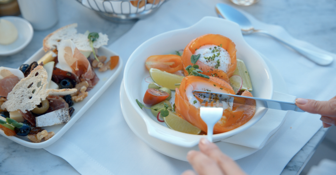 Person cuts into smoked salmon and burrata salad with lime and herbs, beside a platter of cheese, olives, cured meats, and bread.