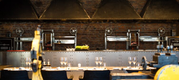 Industrial-style restaurant dining room with brick walls, open-fire grills, metal hoods, and set tables.