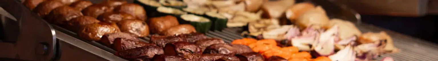 Meat, zucchini, carrots, onions, and bread grilling over glowing coals on a large barbecue grill.