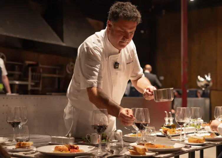 Chef in a white jacket plates dishes at a restaurant counter lined with wine glasses and prepared meals.