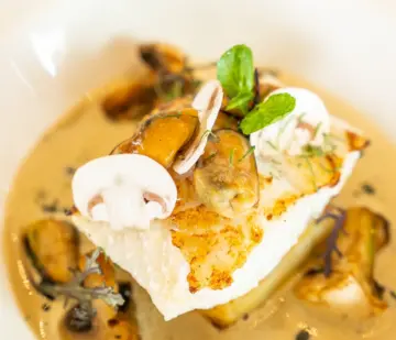 Seared fish fillet with mushrooms, creamy sauce, shaved mushroom, and fresh mint in a white bowl.