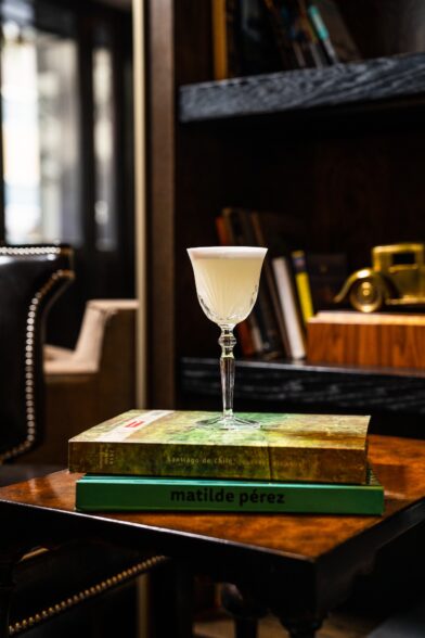 A pale cocktail in a stemmed glass rests on art books atop a wooden table in a dim lounge.