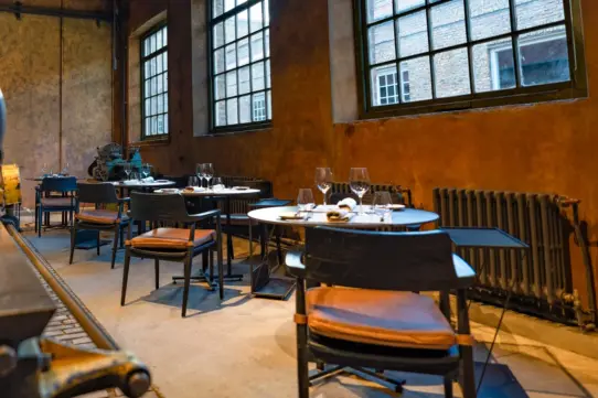 Industrial-style restaurant with set tables, vintage machinery, exposed beams, and tall factory windows letting in daylight.