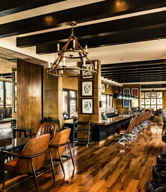 Warm, wood-paneled restaurant interior with leather chairs, a long bar, chandeliers, and framed magazine covers.
