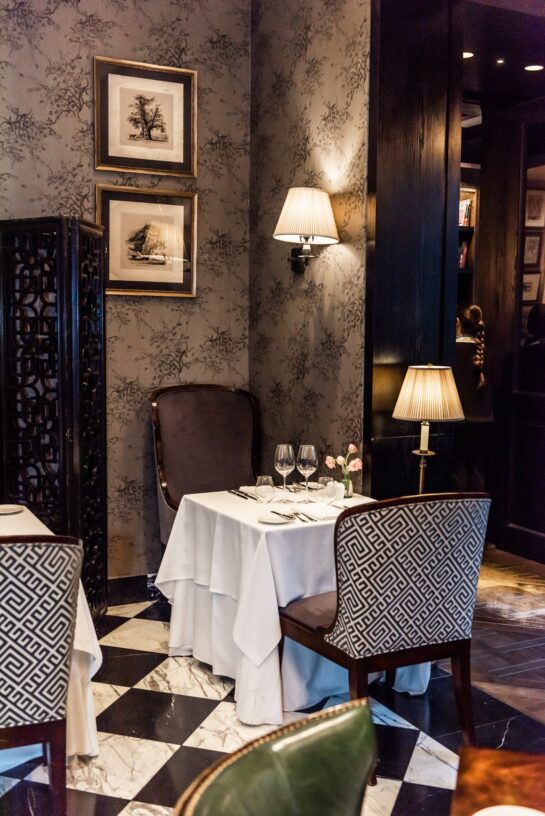 Elegant restaurant dining room with a candlelit table, patterned chairs, framed artwork, and black-and-white marble flooring.