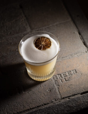 A frothy golden cocktail garnished with a dried orange slice sits on a rustic stone surface.