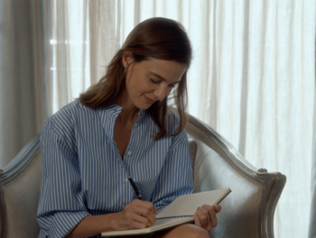 EDITORIAL INTERNA 2 Woman in a blue striped shirt writes in a notebook while seated in an upholstered armchair by a bright window.