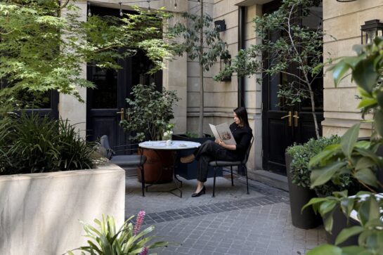 Woman reading a menu at a table in a lush, stone-walled courtyard cafe with hanging string lights and potted plants.