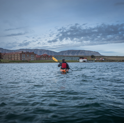 A kayaker paddles across choppy water toward a coastal town beneath cloudy skies and distant mountains.