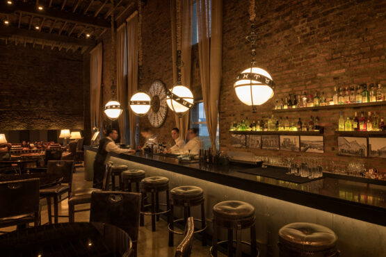 Warm, industrial-style cocktail bar with brick walls, globe pendant lights, leather stools, liquor shelves, and bartenders at work.
