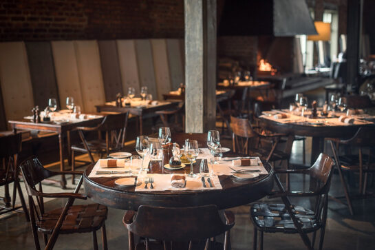 NZ6_6174-EDIT_OPTIMIZADA Comedor de restaurante cálido y rústico con mesas fijas, copas de vino, sillas de madera, paredes de ladrillo y una chimenea resplandeciente.
