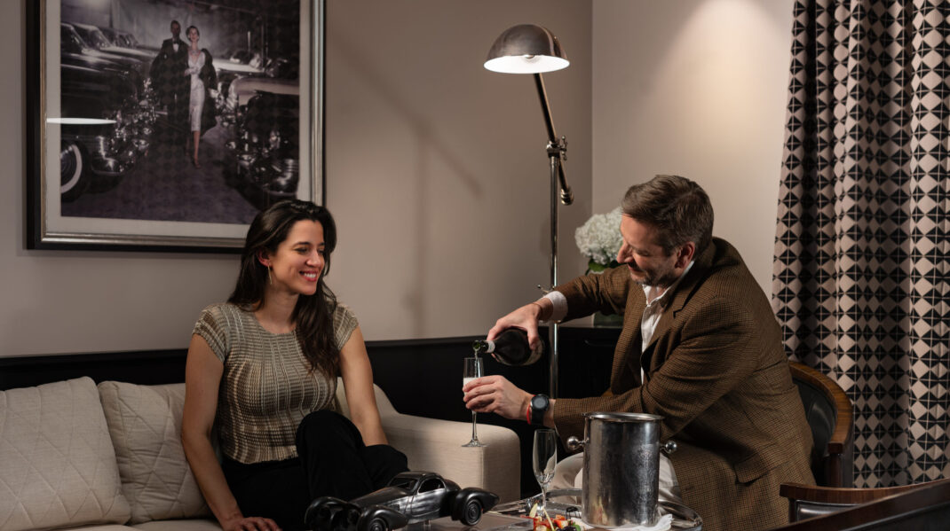A man pours champagne for a smiling woman seated on a sofa in an elegant lounge.
