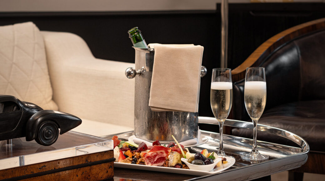 Luxury hotel lounge table with chilled champagne, two flutes, charcuterie, olives, and a cream sofa.