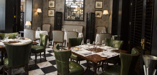 Elegante comedor de restaurante con candelabro de cristal, mesas fijas, sillas de cuero verde, paredes estampadas y suelo de baldosas en blanco y negro.