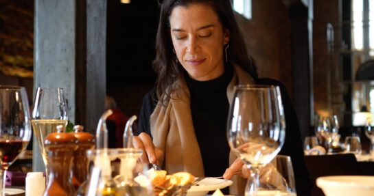 resto3 Mujer cenando en un restaurante, cortando comida junto a copas de vino y una decantador en la mesa.