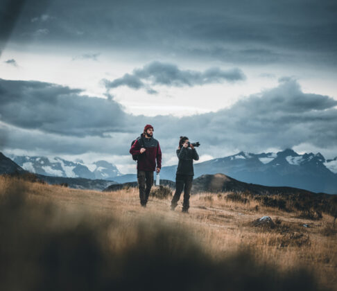 Two hikers cross a golden alpine meadow beneath dramatic storm clouds, with snow-capped mountains in the distance.